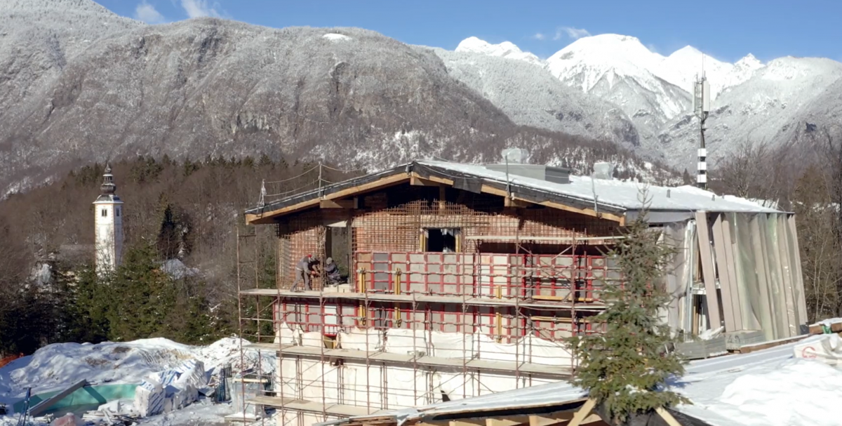 hotel bohinj Stavba v gradnji z odri, postavljena v zasneženo gorsko pokrajino. Gradbišče obdajajo zasneženi vrhovi in drevesa, v ozadju pa je pod jasnim modrim nebom viden cerkveni stolp.