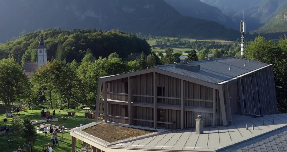 hotel bohinj Med zelenimi drevesi in hribi stoji sodobna lesena stavba s poševno streho, v bližini na travi počivajo ljudje, v ozadju pa se pod jasnim nebom vidi cerkev z visokim stolpom.