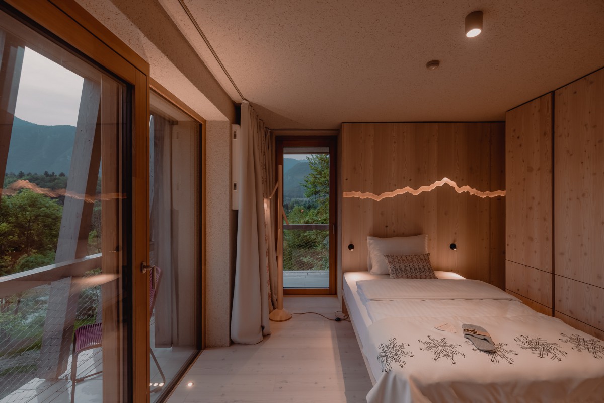 hotel bohinj Udobna spalnica z lesenimi stenami, enojno posteljo z vzorčasto posteljnino, mehko razsvetljavo, velikimi steklenimi vrati in oknom s pogledom na gore in zelenje. Na postelji leži knjiga.