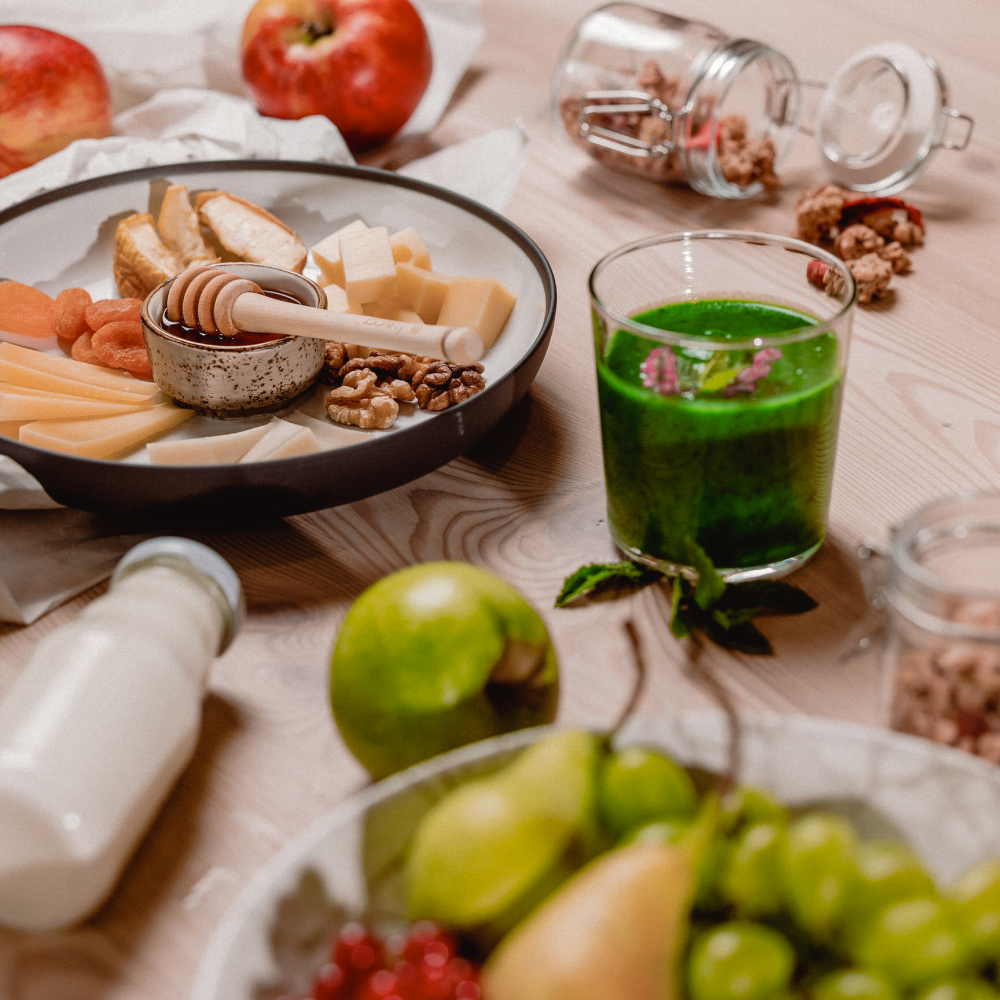 hotel bohinj Miza z narezanim sirom, suhim sadjem, orehi, medom, kozarcem zelenega smoothieja, steklenico mleka, zelenimi jabolki, grozdjem in granatnim jabolkom. Viden je tudi odprt kozarec z orehi in jabolki.