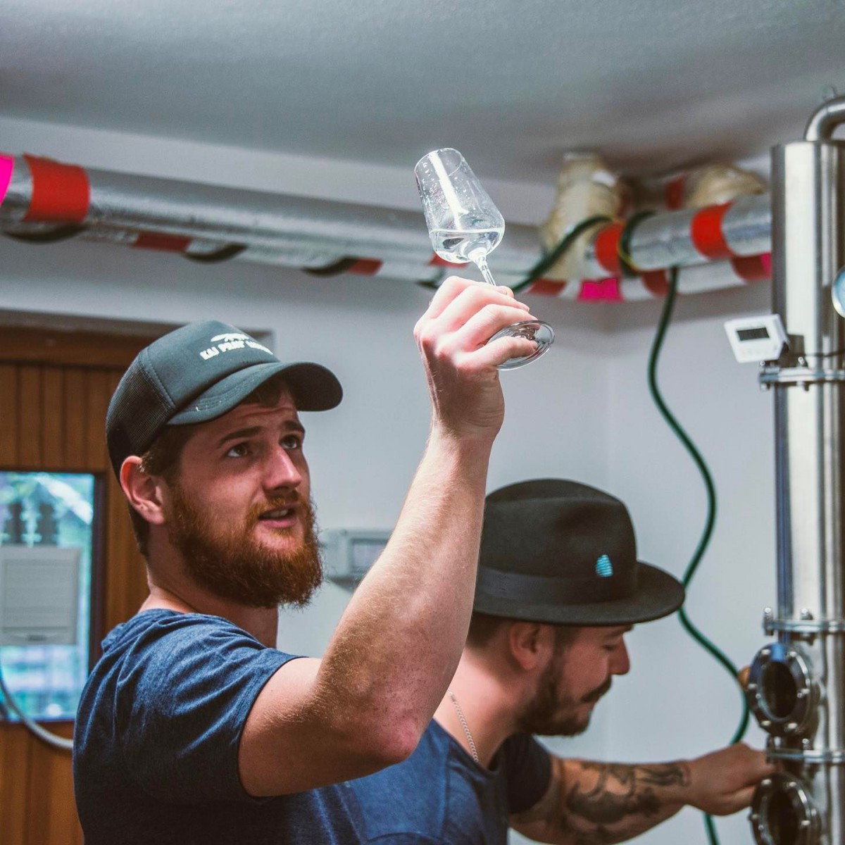 hotel bohinj Bradati moški v kapi drži kozarec in pregleduje njegovo vsebino, medtem ko drugi moški v kapi dela ob industrijski opremi v svetlem prostoru z izpostavljenimi cevmi.