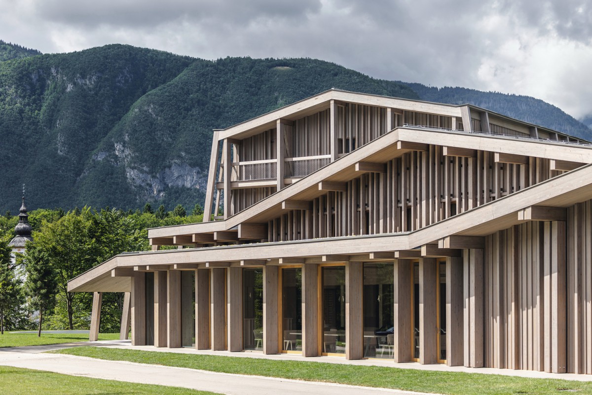 hotel bohinj Na travnatem polju stoji sodobna lesena stavba z velikimi navpičnimi letvami in oglatimi strehami, v ozadju pa so bujno zelene gore in oblačno nebo.