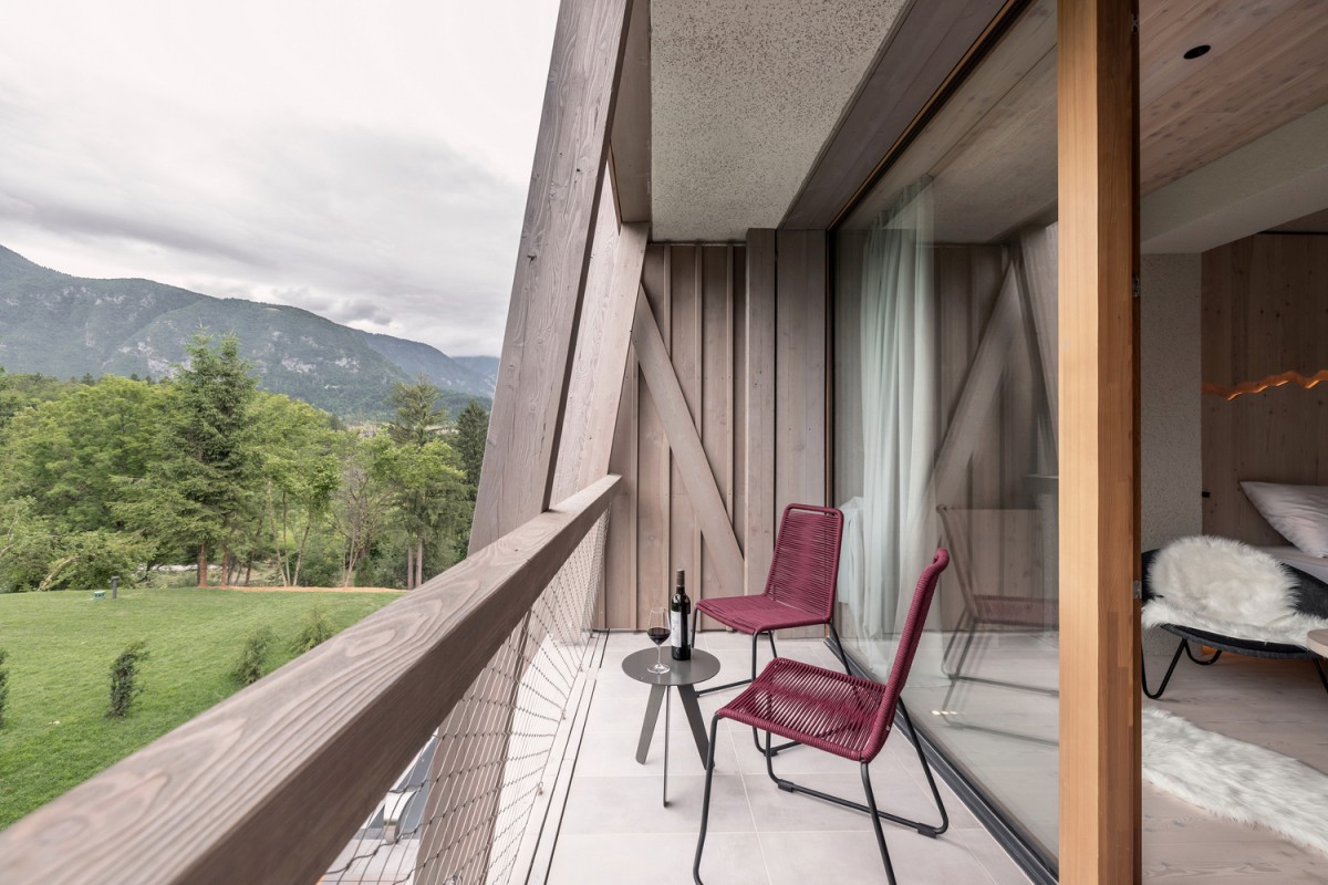 hotel bohinj Sodoben balkon z dvema rdečima stoloma, majhno okroglo mizico s steklenico vina in kozarci, s pogledom na zelena drevesa in oddaljene gore pod oblačnim nebom. Steklena vrata prikazujejo del prijetne spalnice v notranjosti.