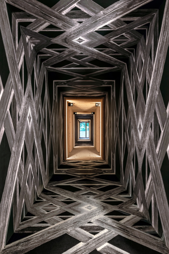 hotel bohinj Hodnik z zapletenimi geometrijskimi vzorci na stenah in tleh ustvarja optično iluzijo, ki vodi do oddaljenih, dobro osvetljenih vrat s pogledom na drevesa in modro nebo zunaj.