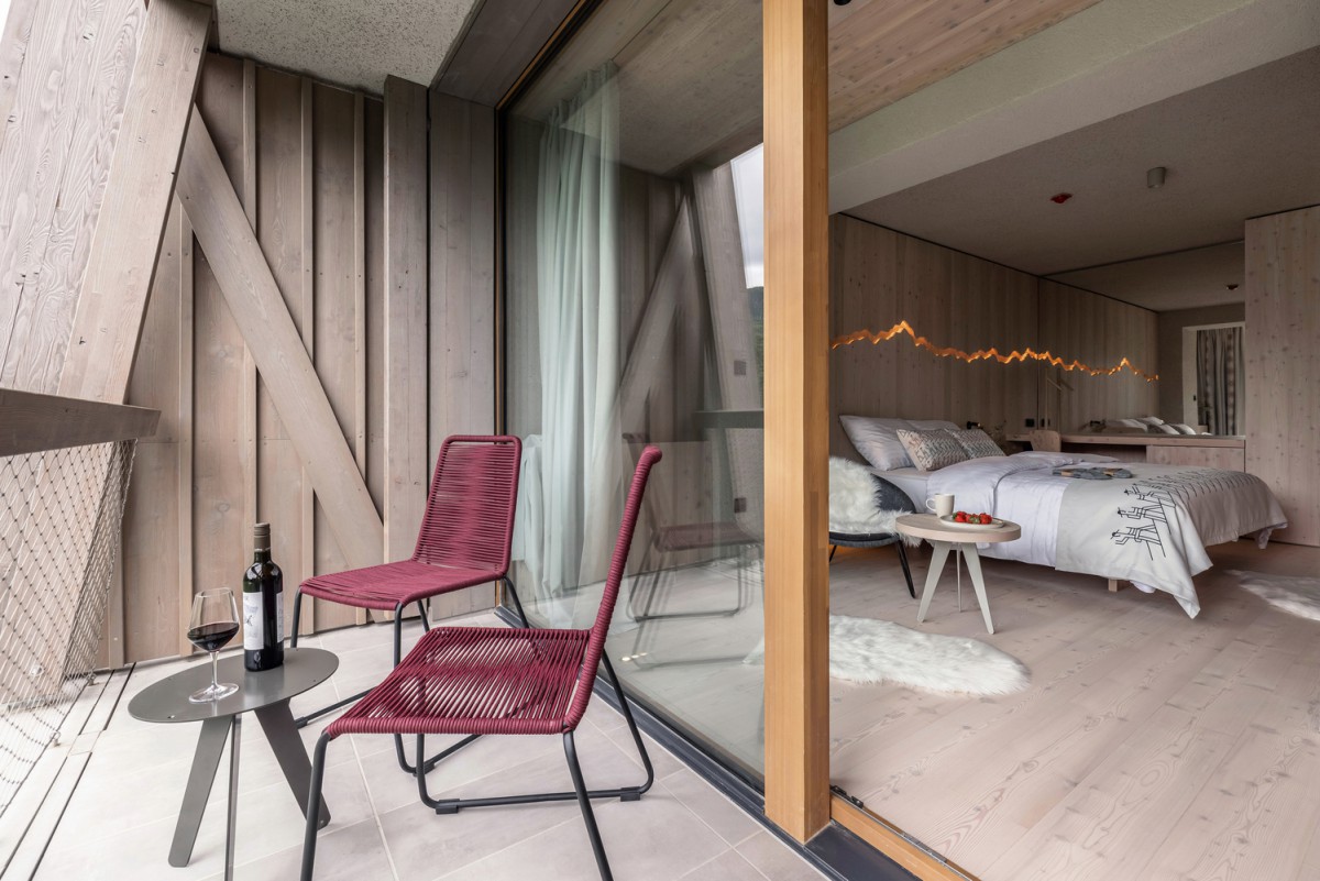 hotel bohinj Sodobna hotelska soba s svetlimi lesenimi tlemi, udobno posteljo in zasebnim balkonom. Na balkonu sta dva rdeča stola, majhna mizica s steklenico vina in kozarci ter pogled na miren zunanji razgled.
