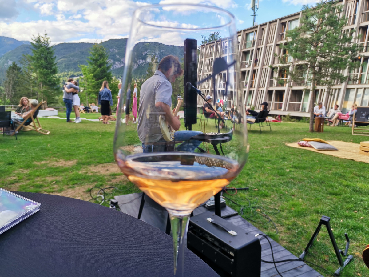 hotel bohinj Na zunanji mizi je v središču pozornosti kozarec z vinom rosé; skozi kozarec je viden glasbenik, ki igra kitaro, na travi se sproščajo ljudje, v ozadju pa so moderna stavba in gore.