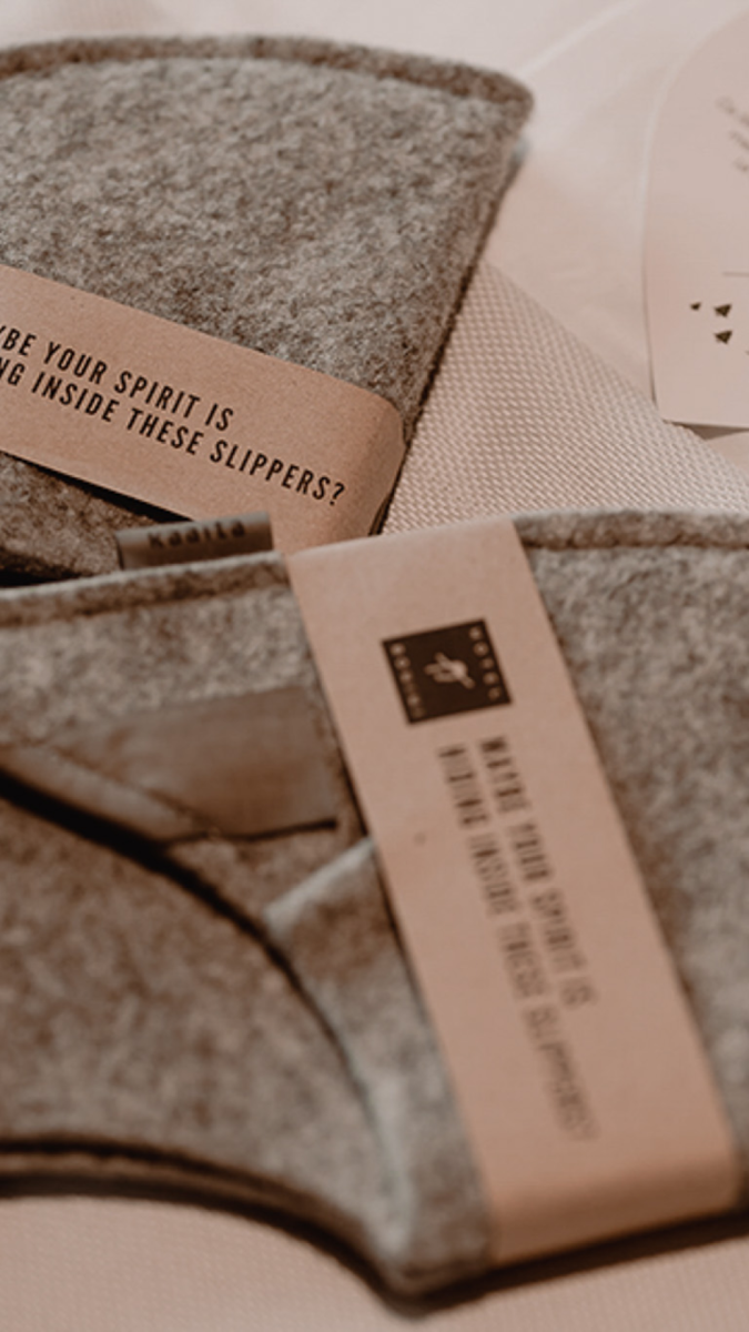 hotel bohinj Close-up of two pairs of grey felt slippers with kraft paper labels, partially visible text, resting on a light-colored fabric surface.