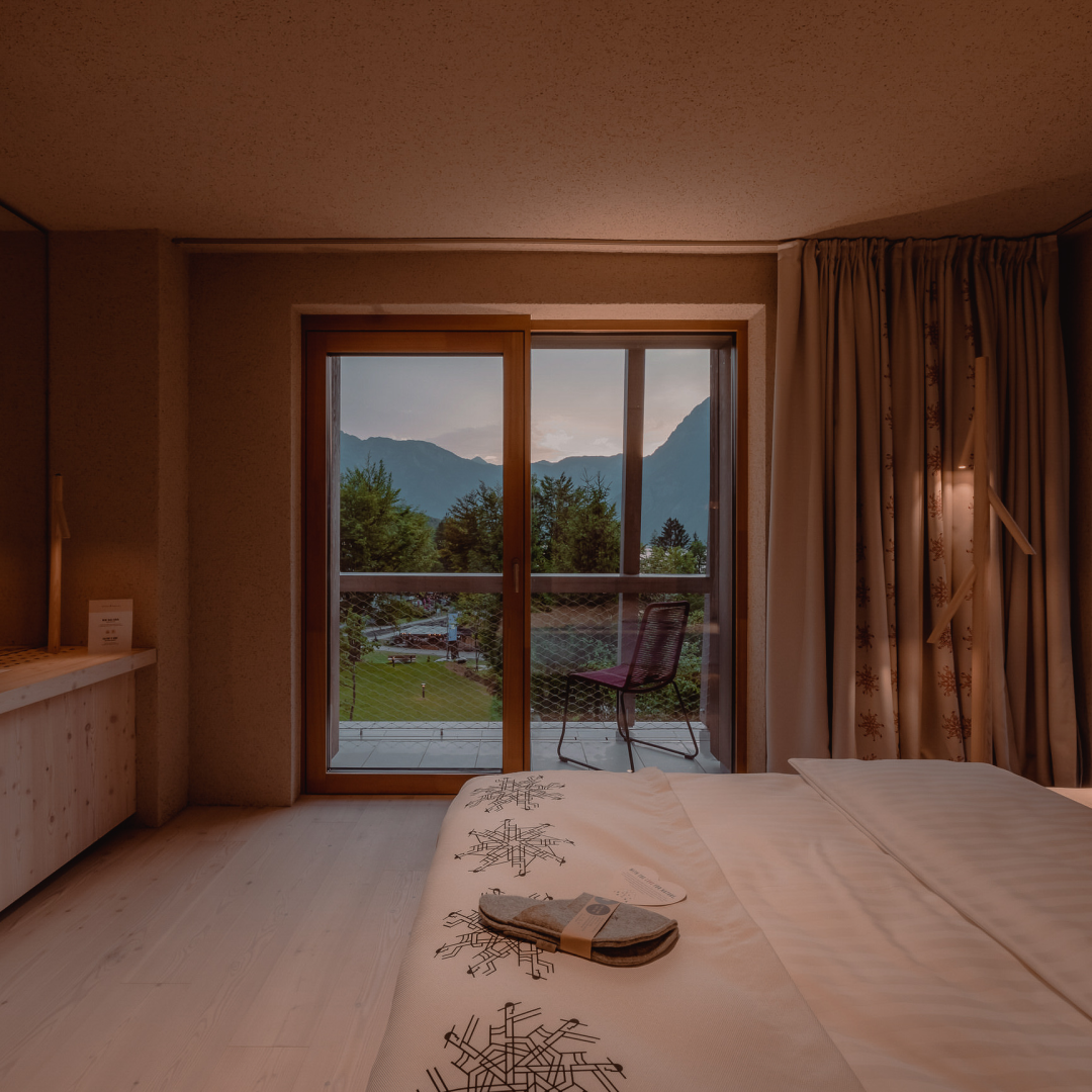 hotel bohinj A cozy hotel room with a neatly made bed, slippers on top, and a balcony door open to a scenic view of mountains, trees, and a soft sunset sky. A chair sits on the balcony.