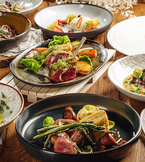 Hotel Bohinj A variety of gourmet dishes are served on a wooden table, including roasted meat with vegetables, lamb chops with herbs, and a selection of side dishes, all artfully presented on elegant plates.