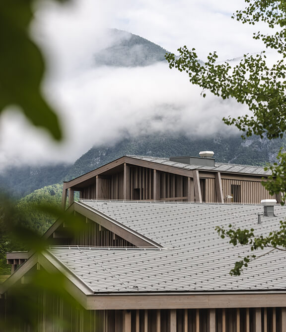 hotel bohinj Sodobno leseno stavbo s sivo streho obdaja zelenje, v ozadju pa so meglene gore in nizko viseči oblaki. V ospredju je prizor uokvirjen z listjem.
