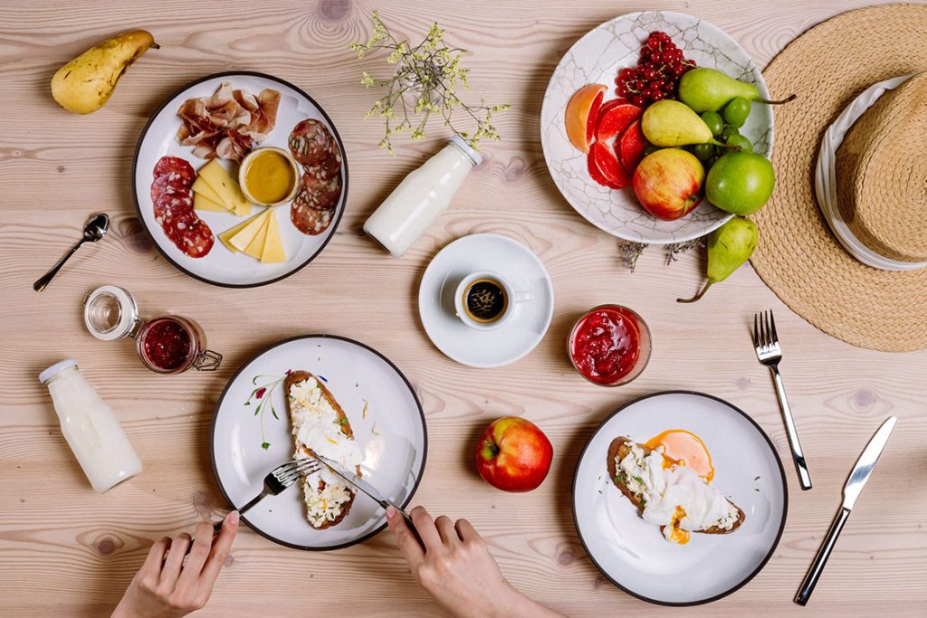 hotel bohinj Na lahki leseni mizi je zajtrk s sadjem, sirom, mesom, jogurtom, marmelado, toastom, skodelico kave, dvema steklenicama mleka in slamnatim klobukom. Dve roki režeta toast na enem krožniku.