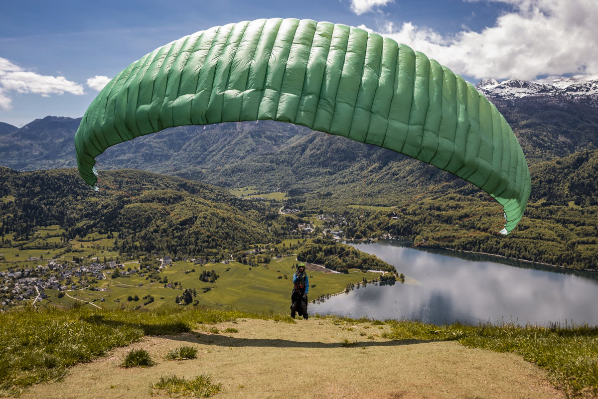 hotel bohinj Oseba se pripravlja na jadralno padalstvo s travnatega pobočja s pogledom na jezero, gozdove in oddaljene gore pod delno oblačnim nebom.