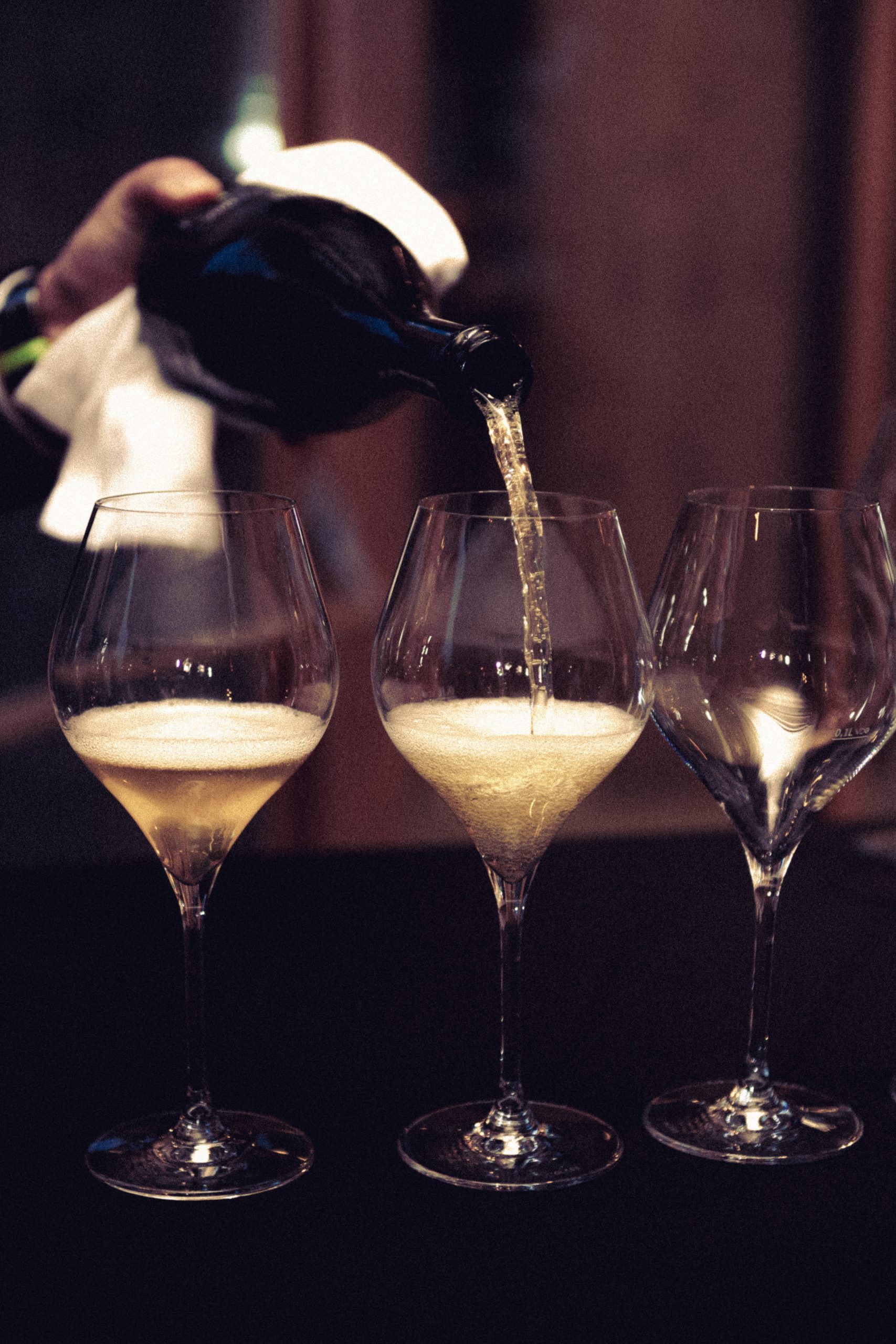 hotel bohinj Oseba iz temne steklenice nalije peneče vino v sredino treh elegantnih kozarcev, pri tem pa si z roko prekrije belo krpo. V dveh kozarcih je vino, eden pa je prazen.