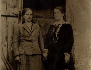 hotel bohinj Dve ženski v staromodnih oblačilih stojita druga ob drugi pred lesenimi vrati in se držita za roke. Zaradi sepije je fotografija zgodovinska, iz začetka 20. stoletja ali konca 19. stoletja.